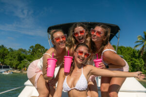 Four women in white swimwear with red sunglasses, holding pink cups, smiling on a boat in Miami under sunny skies.