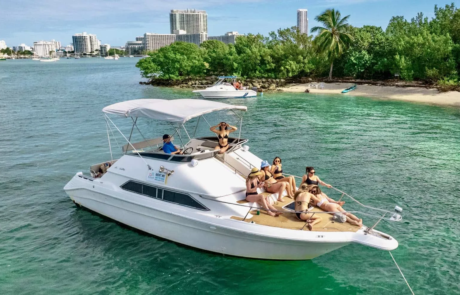 Aerial view of a white boat with a group of people sunbathing, anchored near the shore with Miami city buildings in the background.