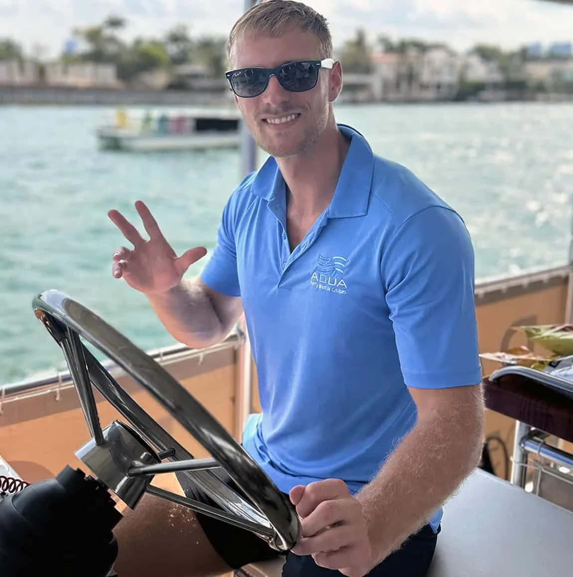 A man wearing a blue polo shirt and sunglasses is steering a boat. He is smiling and giving a thumbs-up gesture. Water and other boats are visible in the background.