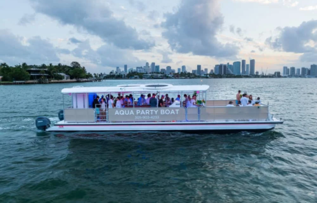 Aqua Party Boat with passengers on board, cruising on the water with a city skyline in the background during dusk. Aqua Party Boat with passengers on board, cruising on the water with a city skyline in the background during dusk.