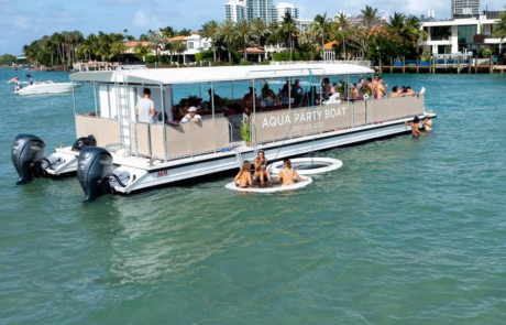 A party boat named “Aqua Party Boat” with people on board and others floating nearby on an inflatable ring, on a sunny day with clear green water and coastal buildings in the background. A party boat named "Aqua Party Boat" with people on board and others floating nearby on an inflatable ring, on a sunny day with clear green water and coastal buildings in the background.