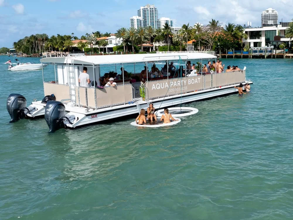 A party boat named "Aqua Party Boat" with people on board and others floating nearby on an inflatable ring, on a sunny day with clear green water and coastal buildings in the background.