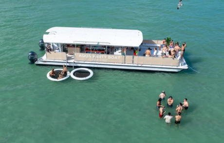 Aerial view of a party boat labeled “Aqua Party Boat” with people onboard and others swimming in turquoise waters. Aerial view of a party boat labeled "Aqua Party Boat" with people onboard and others swimming in turquoise waters.