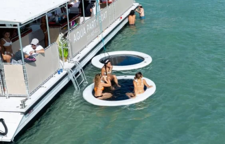 People relax on circular floats in the water next to an “Aqua Party Boat” on a sunny day, with tropical coastline in the background. People relax on circular floats in the water next to an "Aqua Party Boat" on a sunny day, with tropical coastline in the background.