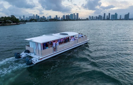 Aerial view of a “Aqua Party Boat” filled with passengers cruising on the water, with a city skyline in the background. Aerial view of a "Aqua Party Boat" filled with passengers cruising on the water, with a city skyline in the background.