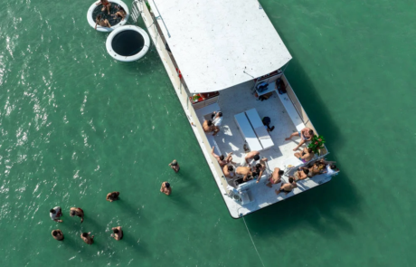 Aerial view of people sunbathing and swimming around a white flatboat in clear green waters, with a smaller boat nearby. Aerial view of people sunbathing and swimming around a white flatboat in clear green waters, with a smaller boat nearby.