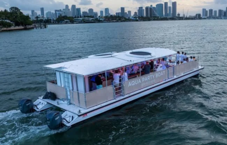 Aerial view of a party boat labeled “Aqua Party Boat” cruising on water with passengers on board, city skyline in the background. Aerial view of a party boat labeled "Aqua Party Boat" cruising on water with passengers on board, city skyline in the background.
