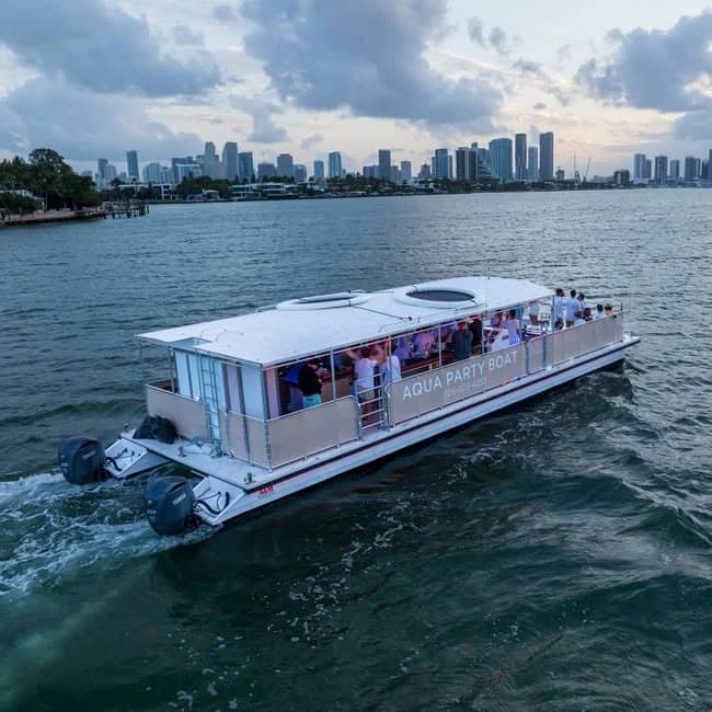Aerial view of a party boat labeled "Aqua Party Boat" cruising on water with passengers on board, city skyline in the background.