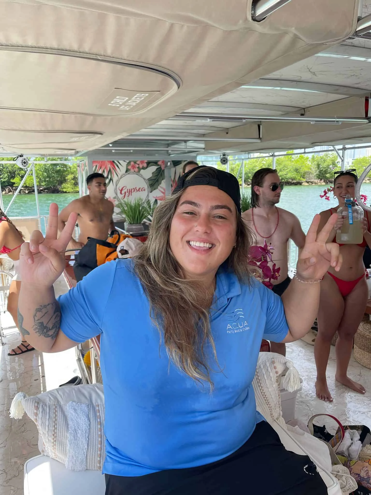 A person in a blue shirt and black cap poses with a peace sign on a boat, with several other individuals in swimwear and a green waterway in the background.